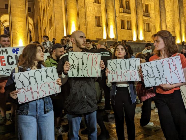 4 protesters carrying white board with Georgian text, in the background more protesters